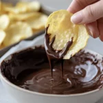 A hand dipping a potato chip into melted chocolate with a tray of prepared chips in the background.