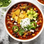 Vegan Chili Recipe with Black Beans in a white bowl, topped with avocado, jalapeño slices, cilantro, tortilla chips, and vegan sour cream.