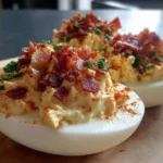 Close-up of creamy Million Dollar Bacon Deviled Eggs topped with crispy bacon, smoked paprika, and fresh chives on a wooden board.
