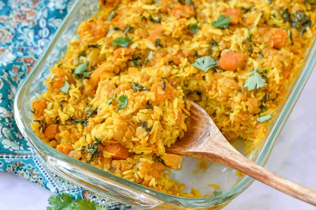 Red Lentil Casserole in a glass baking dish with basmati rice, sweet potato, herbs, and a wooden spoon scooping a serving.