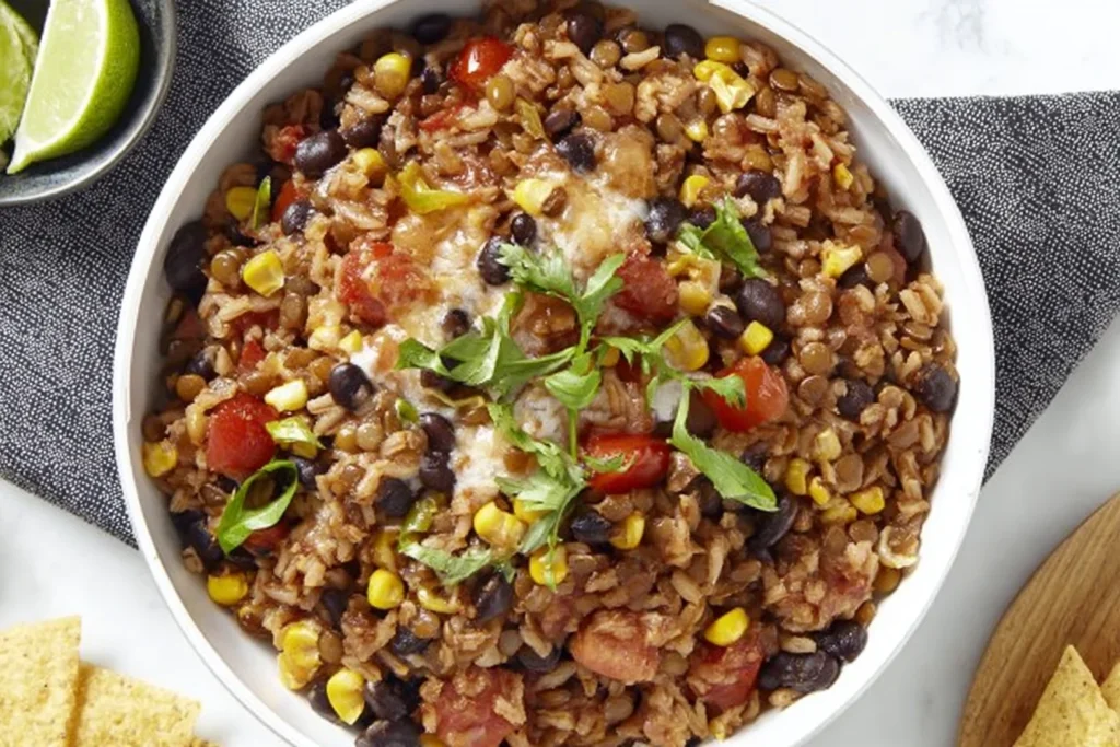Southwest Lentils And Rice Skillet served in a white bowl with black beans, corn, tomatoes, melted cheddar, and cilantro, with lime wedges nearby.