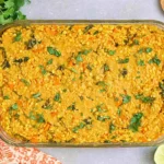 Red Lentil Casserole baked in a glass 9x13 dish, topped with chopped cilantro, with lime wedges and fresh herbs on the side.