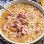 Creamy Ham and Bean Soup with tender navy beans, diced ham, carrots, and herbs in a white bowl with bread on the side.
