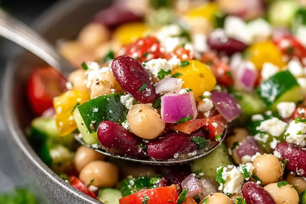 Mediterranean Bean Salad With Feta in a spoon with kidney beans, chickpeas, cucumber, tomatoes, red onion, herbs, and crumbled feta.