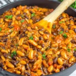Cowboy Beans simmering in a skillet with mixed beans in smoky BBQ tomato sauce, garnished with parsley and stirred with a wooden spoon.