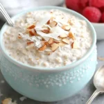 Creamy Coconut Cream Pie Overnight Oats topped with coconut flakes and sliced almonds in a blue bowl with raspberries
