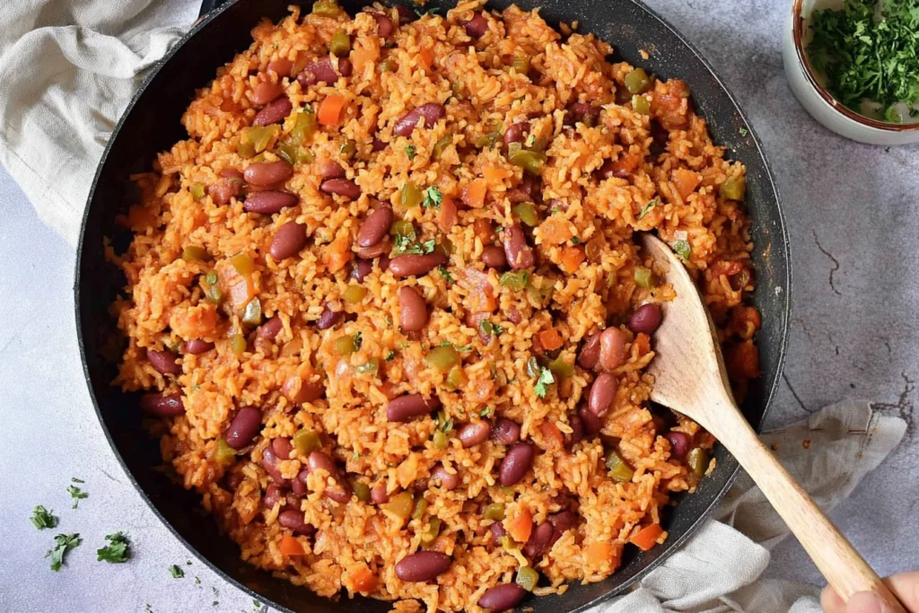 Spanish Rice And Beans cooking in a skillet with fluffy rice, kidney beans, diced peppers, and spices being stirred with a wooden spoon