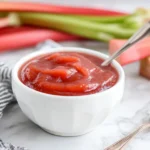 Easy Rhubarb Sauce in a white bowl with spoon and fresh rhubarb stalks
