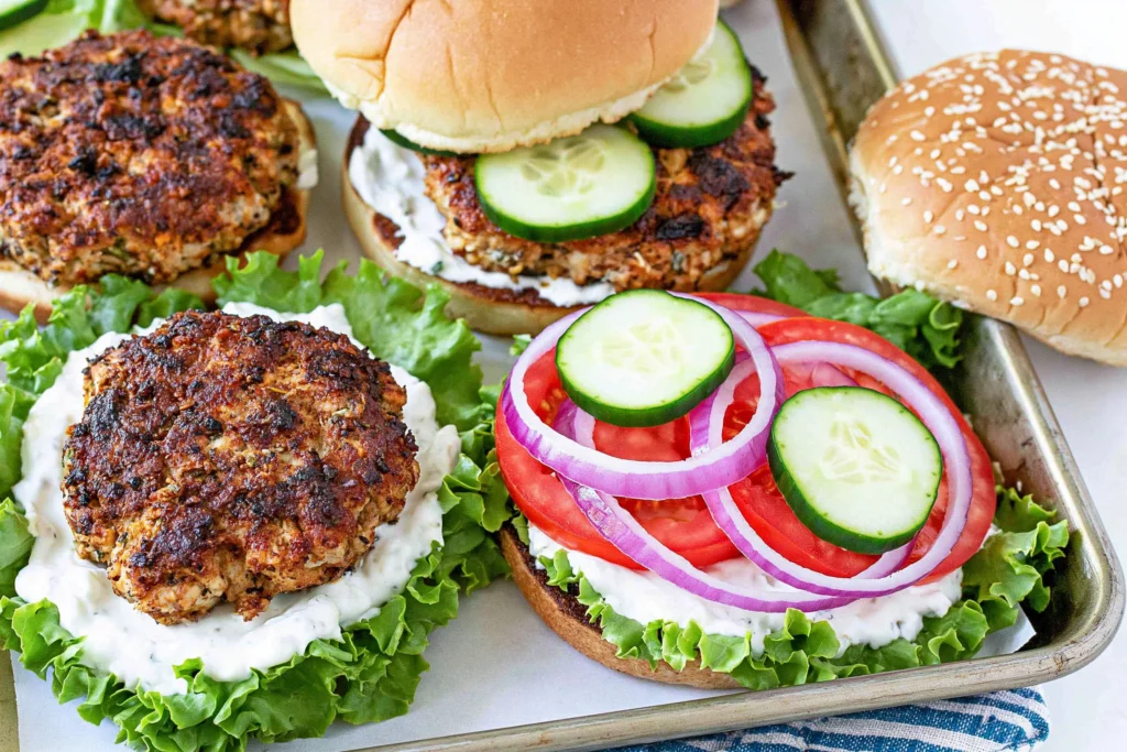 Greek Feta Turkey Burgers being assembled with tzatziki, lettuce, tomato, cucumber, and red onion on buns