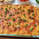 Baked Vegetarian Cheesy White Bean Buffalo Dip in a glass dish topped with melted cheddar and green onions, served with crackers and veggies