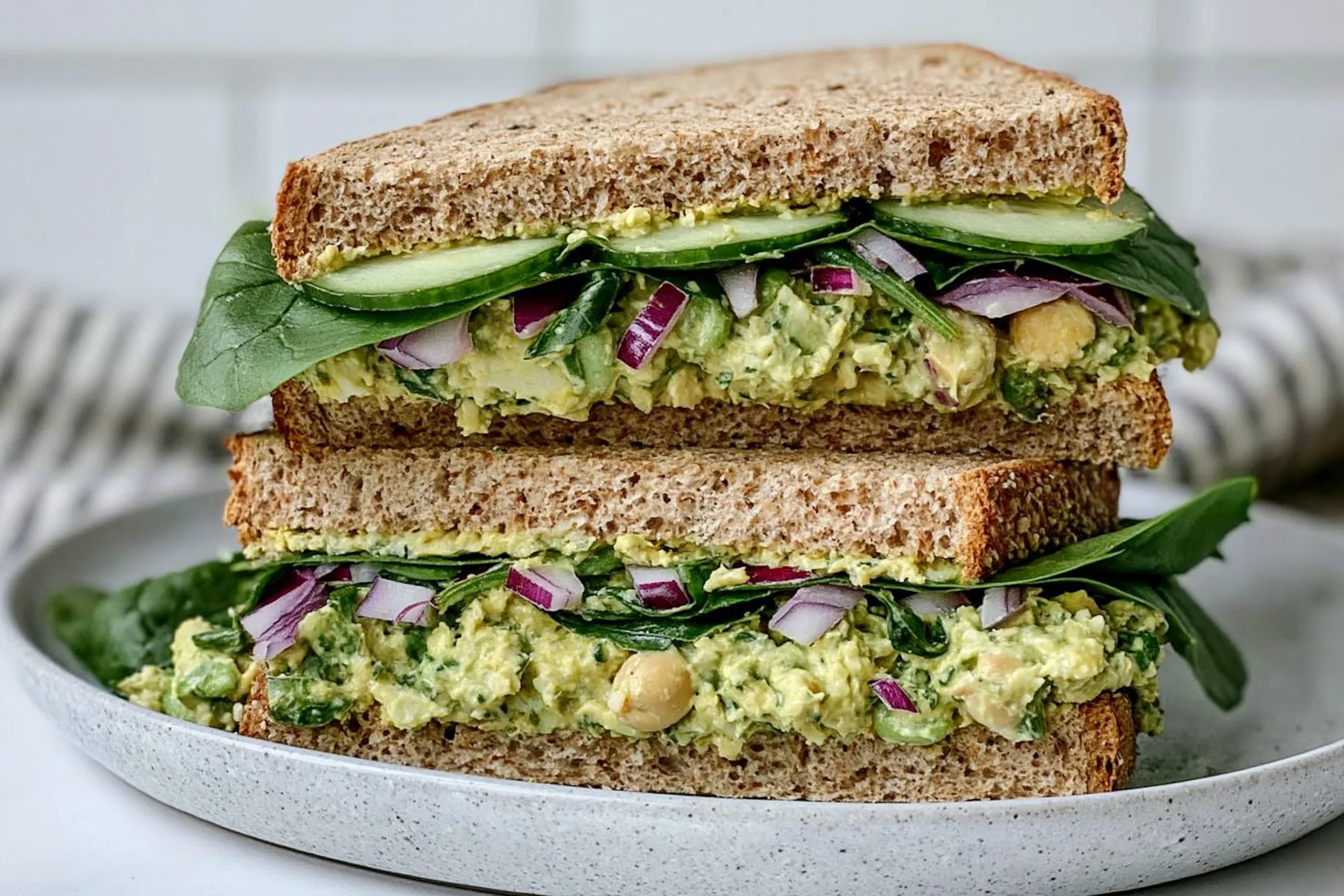 Green Goddess Salad Sandwich with chickpea avocado filling, cucumber, spinach, and whole grain bread stacked on a plate