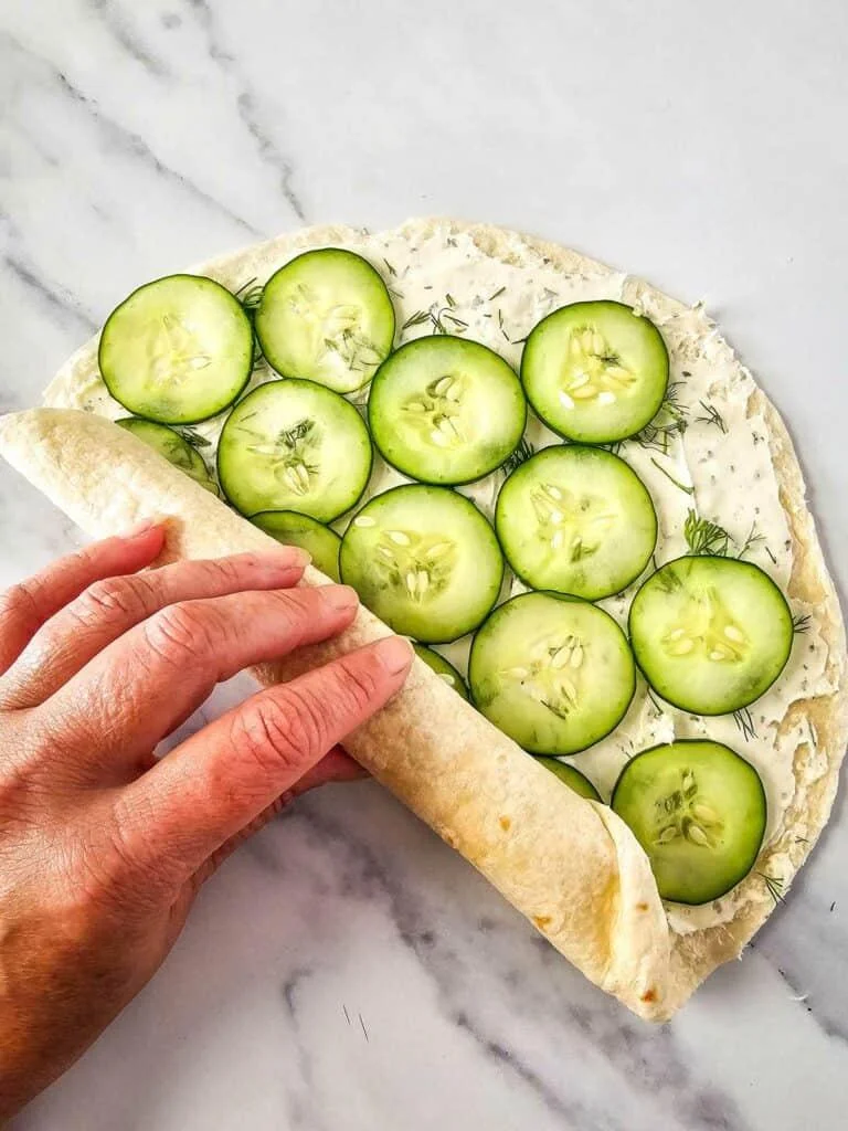 Preparing Cucumber Dill Pinwheels by layering fresh cucumber slices over cream cheese spread on a tortilla