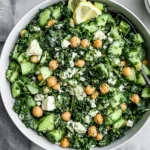Kale Chickpea Salad with Lemon Tahini Dressing with cucumber, chickpeas, feta, fresh herbs, and lemon wedges in a serving bowl.