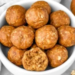 Pumpkin Protein Balls in a white bowl with oats, featuring one protein ball cut open to show its soft, chewy center.