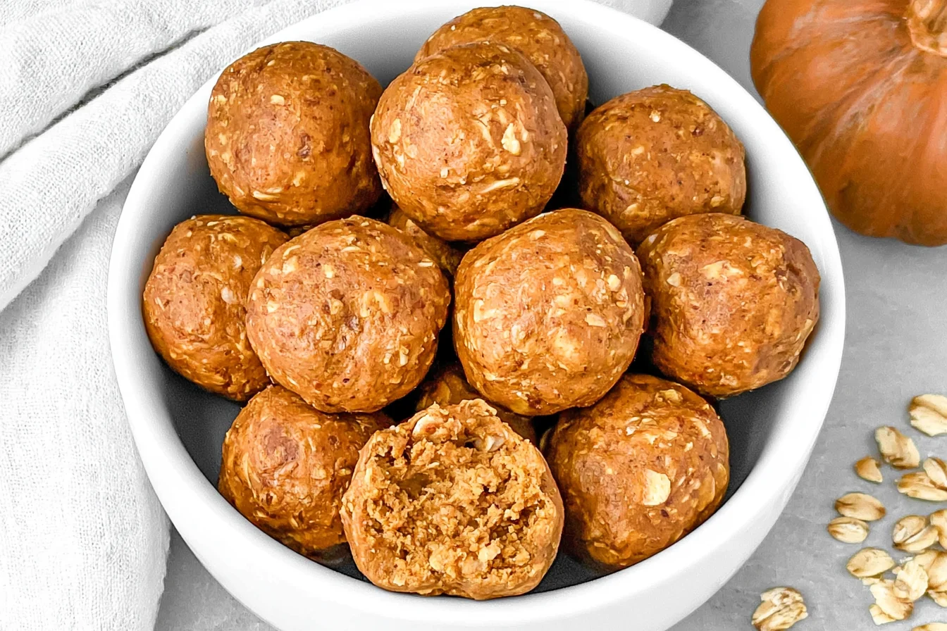 Pumpkin Protein Balls in a white bowl with oats, featuring one protein ball cut open to show its soft, chewy center.