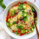 Spring Roll Salad with Spicy Ginger Dressing featuring thin rice noodles, shredded carrots, red bell peppers, cucumber, fresh cilantro, sliced green jalapeños, and roasted peanuts in a white bowl.