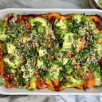 Lightened-Up Salmon Sushi Bake topped with avocado, scallions, edamame, sesame seeds, and spicy sauce in a white casserole dish