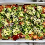 Lightened-Up Salmon Sushi Bake topped with avocado, scallions, edamame, sesame seeds, and spicy sauce in a white baking dish