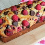 Raspberry Dark Chocolate Banana Bread loaf topped with fresh raspberries and dark chocolate chunks, featuring a golden-brown crust on a wooden board.