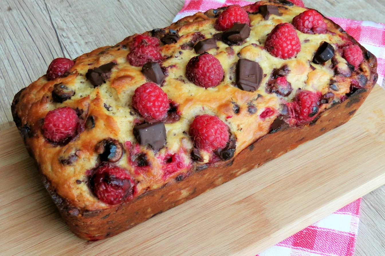 Raspberry Dark Chocolate Banana Bread loaf topped with fresh raspberries and dark chocolate chunks, featuring a golden-brown crust on a wooden board.